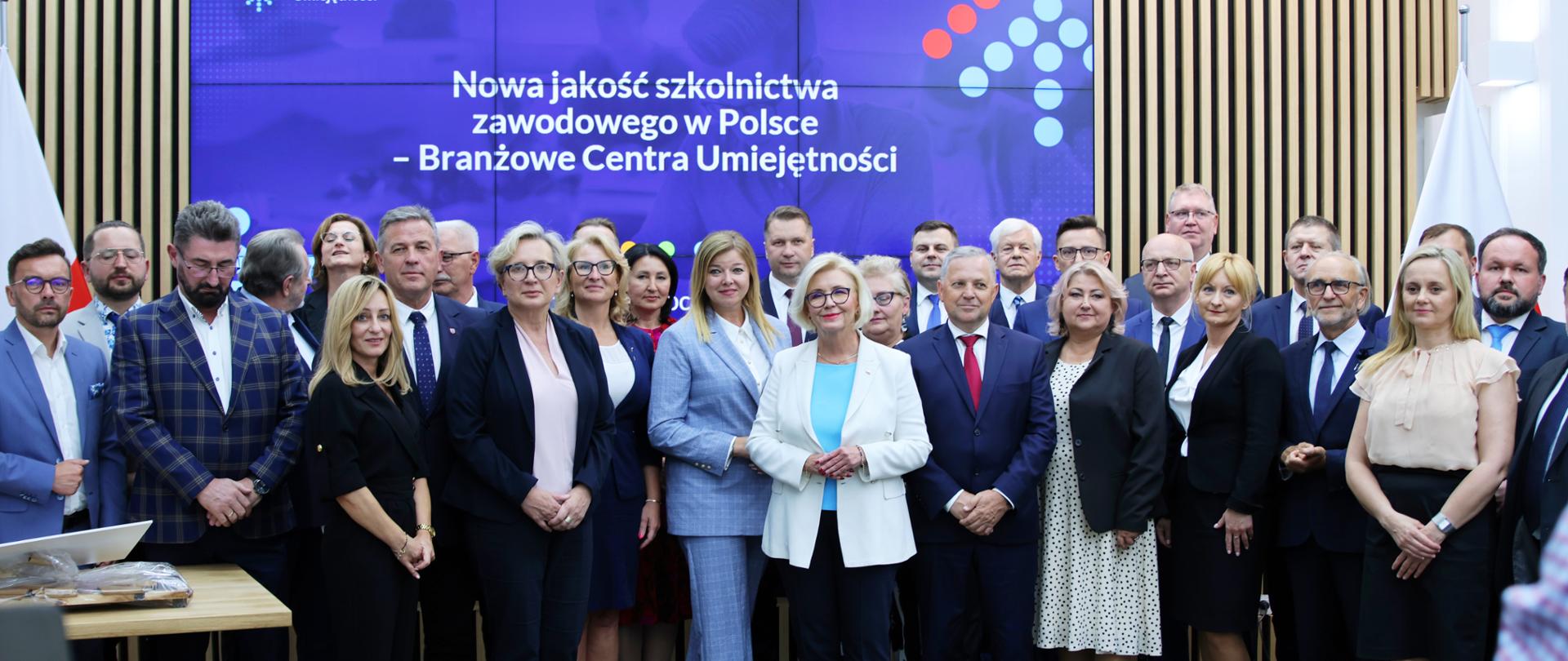 Grupa osób pozujących do zdjęcia, w tle błękitny ekran z napisem Nowa jakość szkolnictwa zawodowego w Polsce – podpisanie pierwszych umów w ramach Branżowych Centrów Umiejętności
