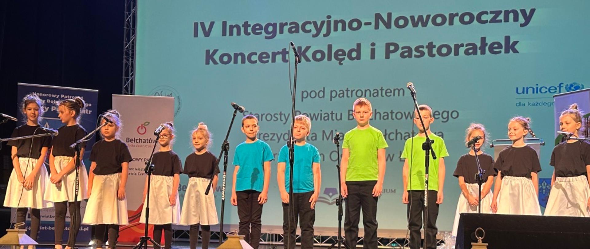 Zdjęcie przedstawia scenę na sali koncertowej MCK PGE GIGANTY MOCY. Na scenie stoi grupa 12 dzieci - kolorowo ubranych. Są to uczniowie Państwowej Szkoły Muzycznej I stopnia w Bełchatowie, którzy wzięli udział w IV Integracyjno-Noworocznym Koncercie Kolęd i Pastorałek w Bełchatowie. W tle za sceną duży wyświetlany napis o treści "IV Integracyjno-Noworoczny Koncert Kolęd i Pastorałek"