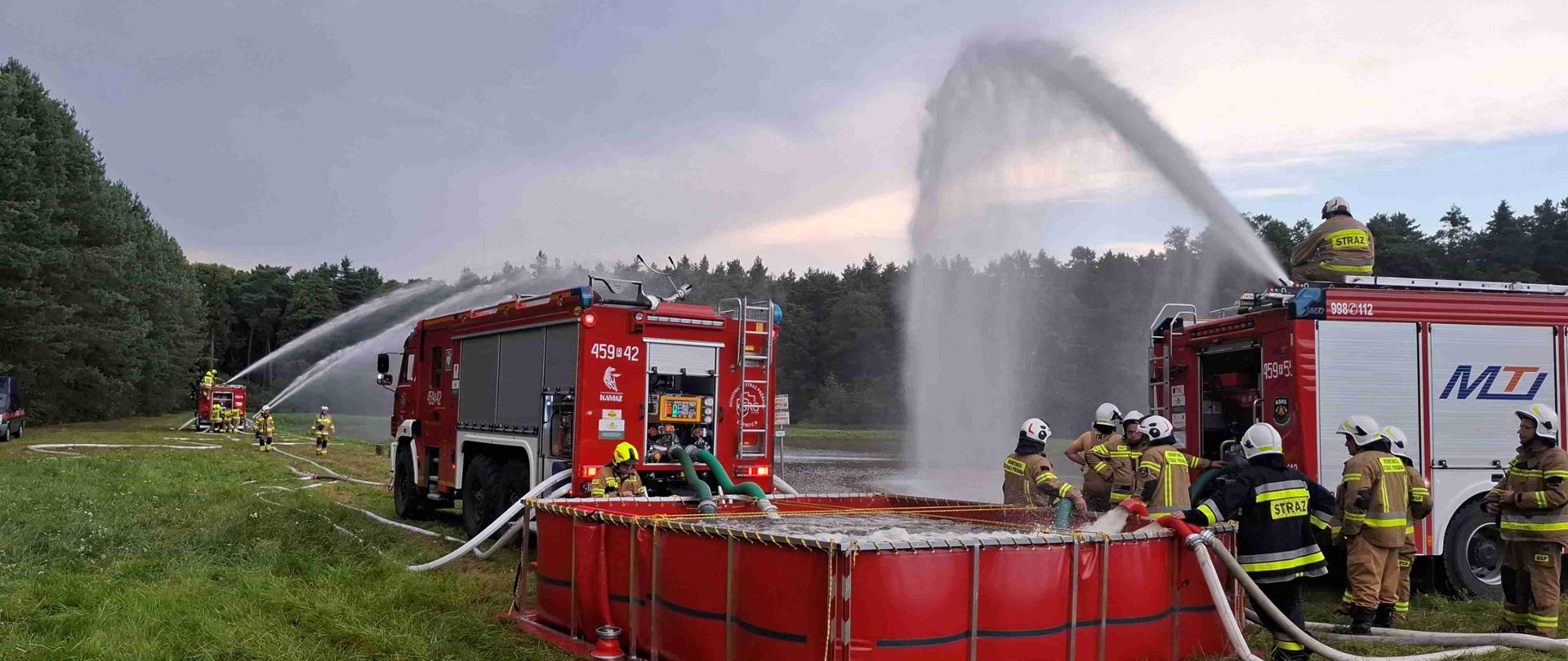 Na pierwszym planie rozłożony duży czerwony zbiornik pełny wody. Do zbiornika jednymi wężami wlewana jest woda, drugimi woda czerpana jest do dwóch dużych samochodów strażackich - jeden z nich z działka na dachu leje wodę do zbiornika. W tle inny samochód który z działka na dachu też leje wodę. Pomiędzy samochodami rozwinięte węże strażackie. W tle las.