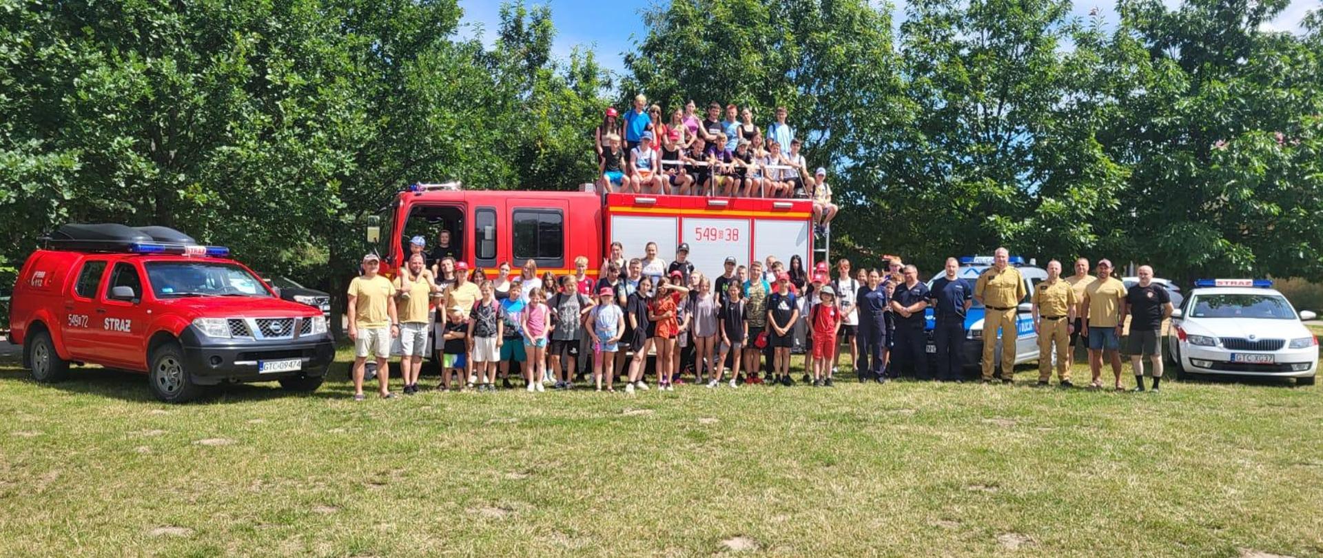 Zdjęcie grupowe uczestników obozu - dużo dzieci, funkcjonariusze policji i policji, część dzieci siedzi na dachu wozu strażackiego. Stoją też radiowóz i dwa samochody strażackie osobowe.