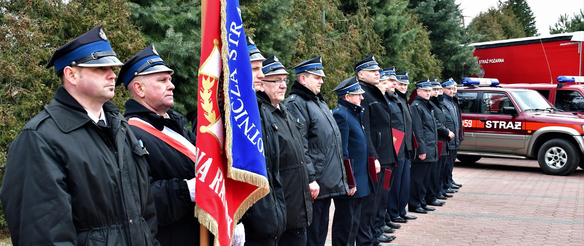 Druhowie Ochotniczych Straży Pożarnych ustawieni w rzędzie, każdy z nich trzyma otrzymaną promesę na zakup samochodów pożarniczych. Na czele rzędu stoi sztandar. W tle widać samochody pożarnicze.