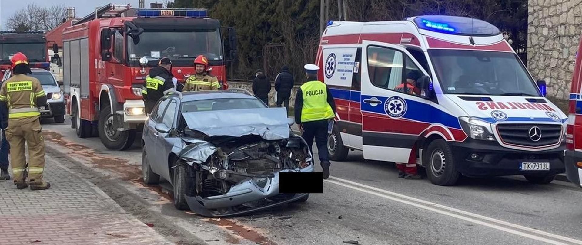 Zdjęcie przedstawia uszkodzony pojazd osobowy. Obok pojazdu znajduje się karetka PRM. Przy pojeździe widać strażaków oraz policjanta. Za ww. pojazdem znajdują się trzy samochody pożarnicze