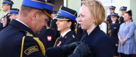 Strażak oficer w mundurze galowym przypina medal kobiecie. W tle goście uroczystości, w dali budynek strażnicy