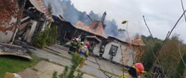Zdjęcie przedstawia zniszczone dwa budynki po pożarze, po prawej stronie budynek mieszkalny jednorodzinny, po lewej stronie budynek garażowo-gospodarczy. Zapadnięte dachy, okopcone ściany, unoszący się dym. Strażacy dogaszają budynek mieszkalny.