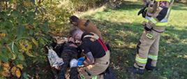 Akcja ratunkowa w terenie zielonym. Na ziemi, pod krzewem, leży poszkodowana osoba opatulona folią termiczną NRC, z opatrunkiem na kolanie. Dwóch strażaków klęczy przy poszkodowanym, udzielając mu pierwszej pomocy, a trzeci strażak stoi obok i nadaje komunikat przez krótkofalówkę.
