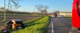 Na zdjęciu widoczne rozbite auto znajdujące się w przydrożnym rowie, W pojeździe za pomocą narzędzi hydraulicznych usunięto lewą stronę karoserii. Obok pojazdu w kolorze miedzi leży ekwipunek w postacie przedmiotów letniego wypoczynku. Na drodze w odległości kilkunastu metrów stoi drugi pojazd uczestniczący w zdarzeniu. Srebrna Skoda Fabia z rozbitym przodem. Na jezdni duża ilość elementów karoserii samochodowej. Pojazdy pożarnicze ustawione w poprzek drogi blokuję ruch w obu kierunkach, 