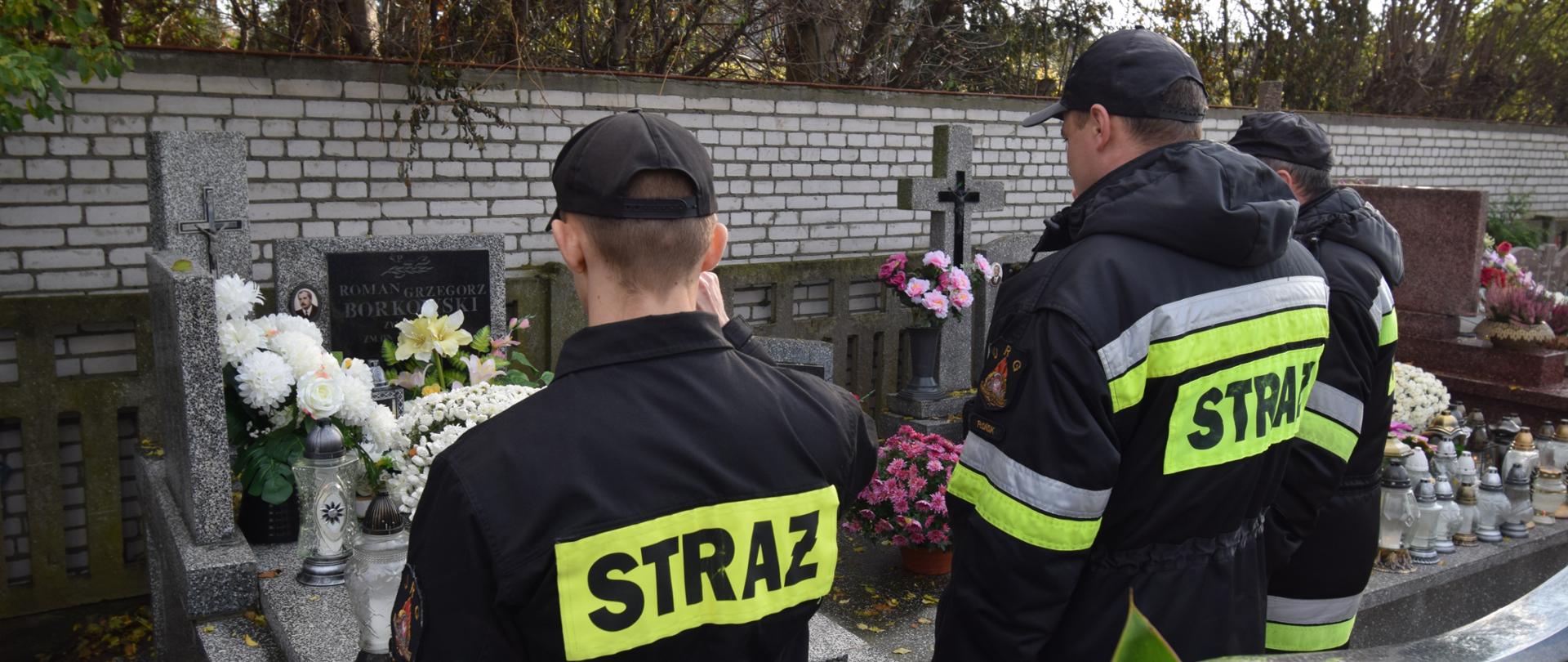 Cmentarz, przy grobie stoi trzech strażaków w ubraniach koszarowych w kolorze czarnym z napisem straż i salutuje. 