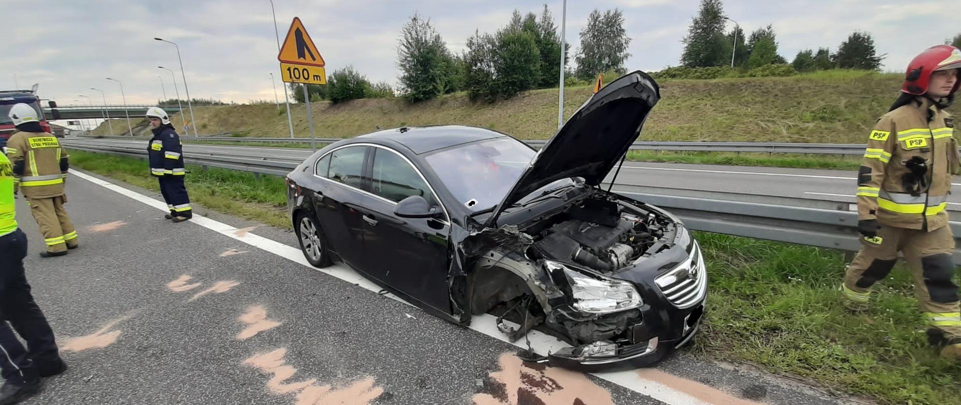 Zdjęcie obrazuje samochód osobowy Opel Insignia po kolizji drogowej. Od samochodu odpadło przednie prawe koło. Jezdnia natomiast jest posypana sorbentem , aby zneutralizować wycieki olejowe. Obok pojazdu przemieszczają się strażacy PSP i OSP. Stoi też policjant. W tle widać infrastrukturę drogową.