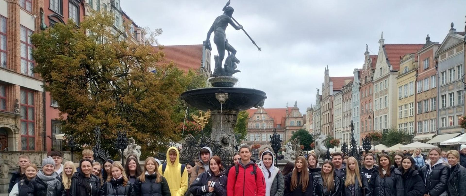 Na zdjęciu grupa uczniów pozuje wspólnie przed fontanną Neptuna na gdańskiej starówce. W tle widać kolorowe kamienice o charakterystycznej architekturze oraz drzewa z jesiennymi liśćmi. To pamiątkowa fotografia ze spaceru po centrum Gdańska.