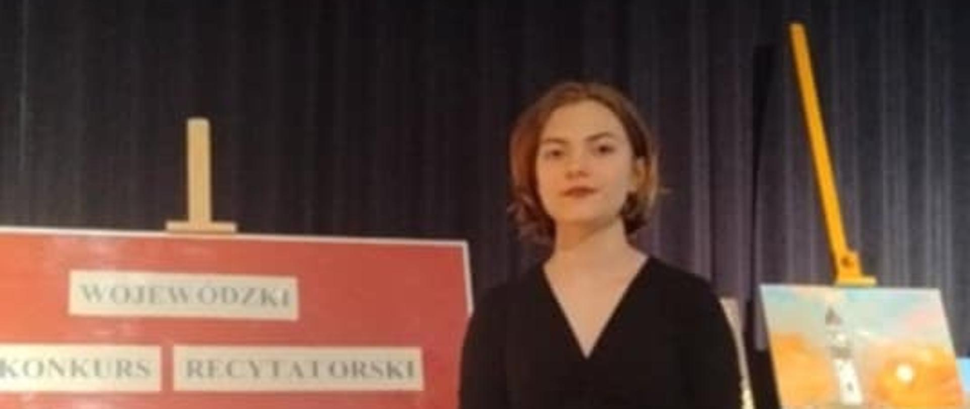 Maria Szymczyk stoi przed malarskimi sztalugami w sali. Ubrana jest w czarną bluzkę i brązowe spodnie wysokie w stanie. Na jednej sztaludze znajduje się napis: "Wojewódzki Konkurs Recytatorski Poezji i Prozy Absolwentów III LO". Na drugiej sztaludze umieszczony jest obraz, pejzaż przedstawiający morze, latarnię morską o wschodzie słońca.