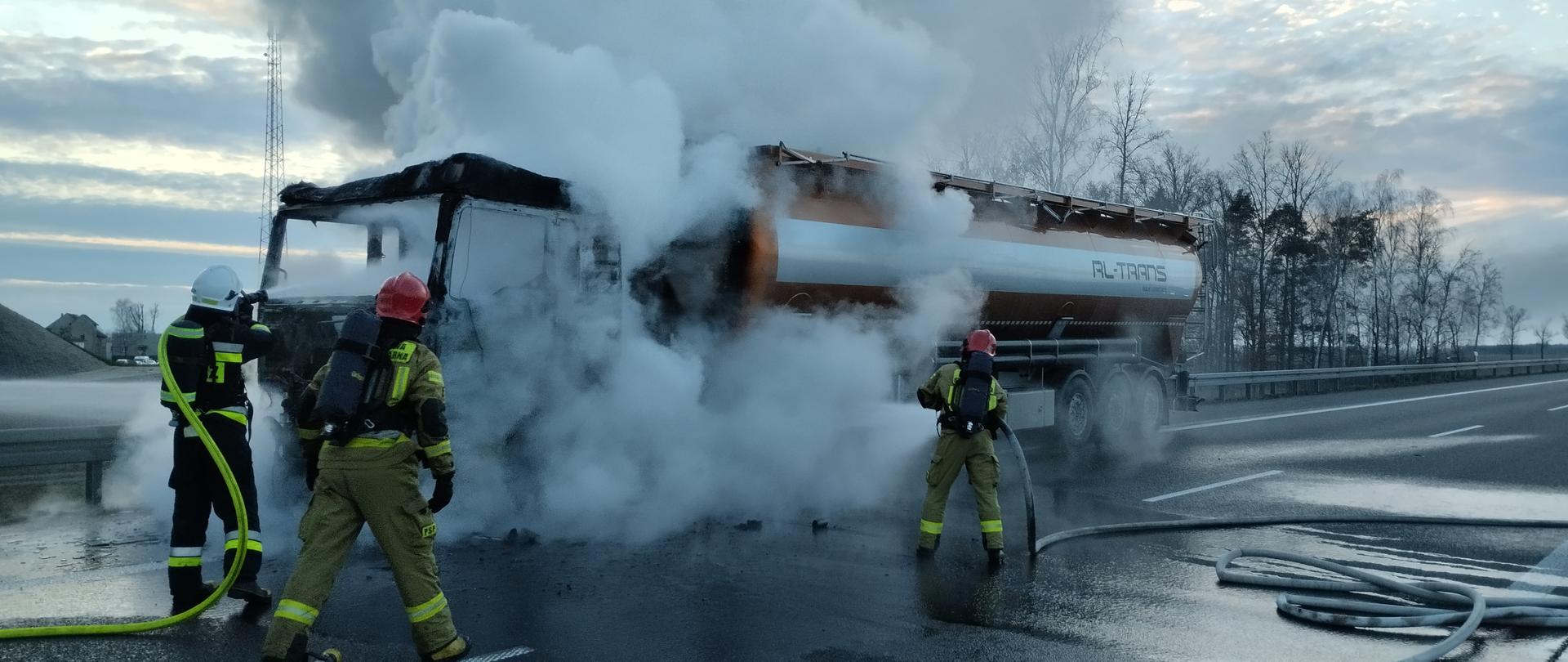 Pochmurny dzień. samochód ciężarowy stojący na drodze z cysterną cały w białym dymie. Obok niej strażacy w jasnych i ciemnych mundurach w czerwonych i białych hełmach trzymają żółte i białe węże strażackie. Na plecach mają ciemne butle apartów oddechowych z powietrzem.