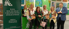 Sześć osób stoi przy stoisku informacyjnym Kasy Rolniczego Ubezpieczenia Społecznego. Stoisko jest ozdobione zielonymi banerami z logo KRUS i hasłem „Pracuj bezpiecznie”. Na stolikach ułożone są ulotki i broszury informacyjne. W tle widoczna jest wystawa kolorowych obrazów o tematyce przyrodniczej.