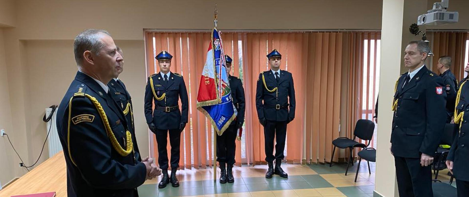 na sali widać od lewej strony umundurowanego komendanta powiatowego z zastępcą, stojących bokiem, potem w centrum widać umundurowany poczet sztandarowy stojący frontem do kadru w centralnej części zdjęcia, kolejno po prawej stronie stoją umundurowani funkcjonariusze.