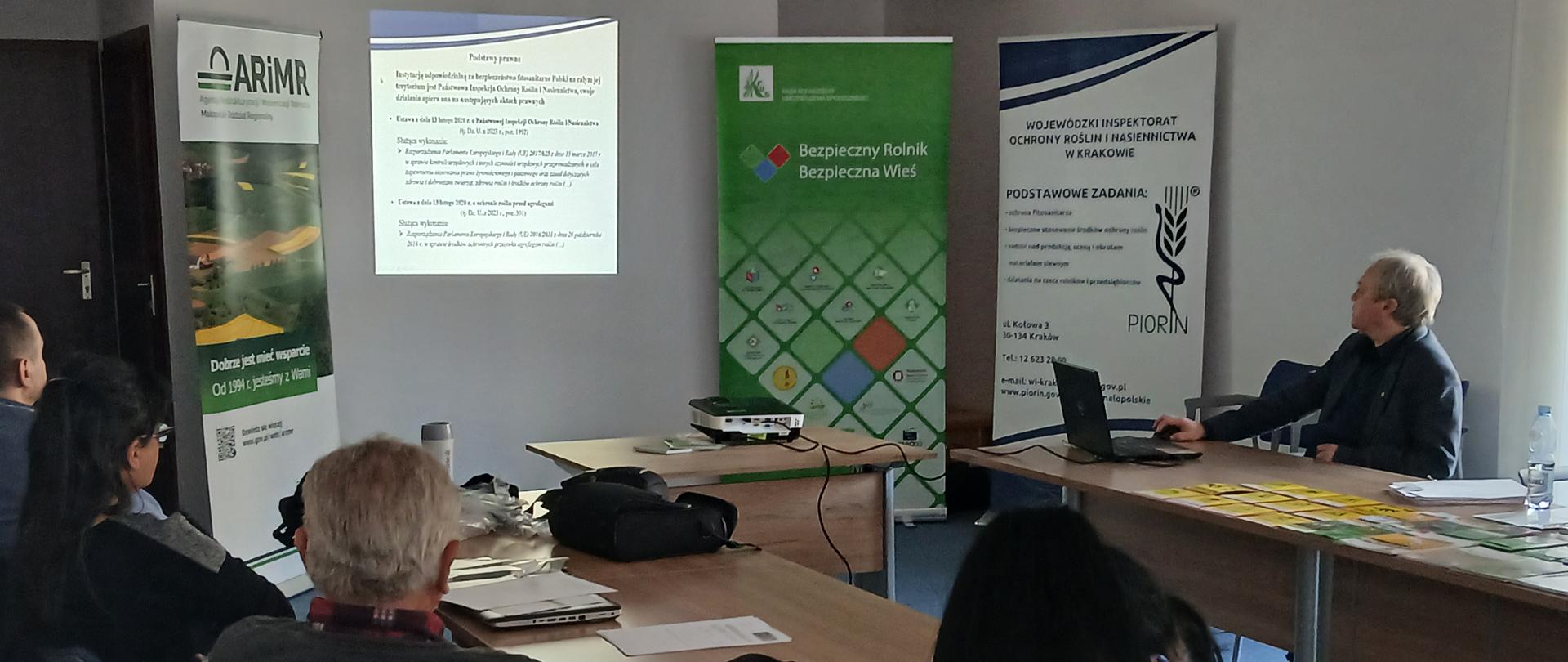 Widok sali szkoleniowej, kilku osób uczestniczących w szkolenie, siedzących przy stole, na drugim planie mężczyzna przeprowadzający szkolenie