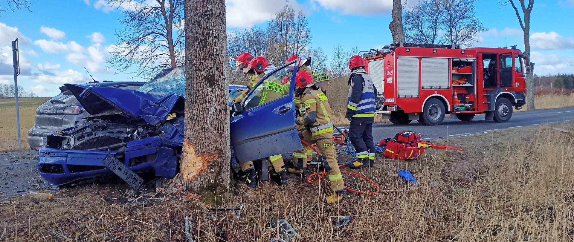 Zdjęcie przedstawia samochód osobowy po uderzeniu w przydrożne drzewo. Strażacy ratownicy wykonują dostęp do osoby poszkodowanej przy użyciu hydrauliki