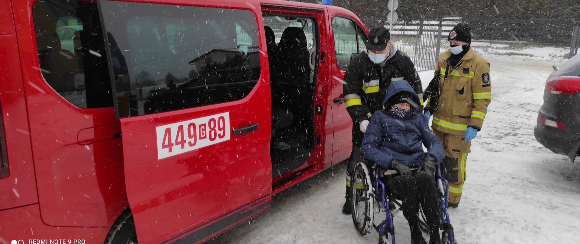 Zdjęcie przedstawia strażaka w ubraniu specjalnym pomagającego poruszać starszej osobie na wózku inwalidzkim