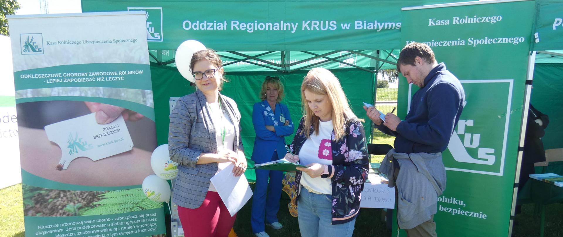 Trzy kobiety i mężczyzna przy stoisku pod namiotem z logo KRUS.