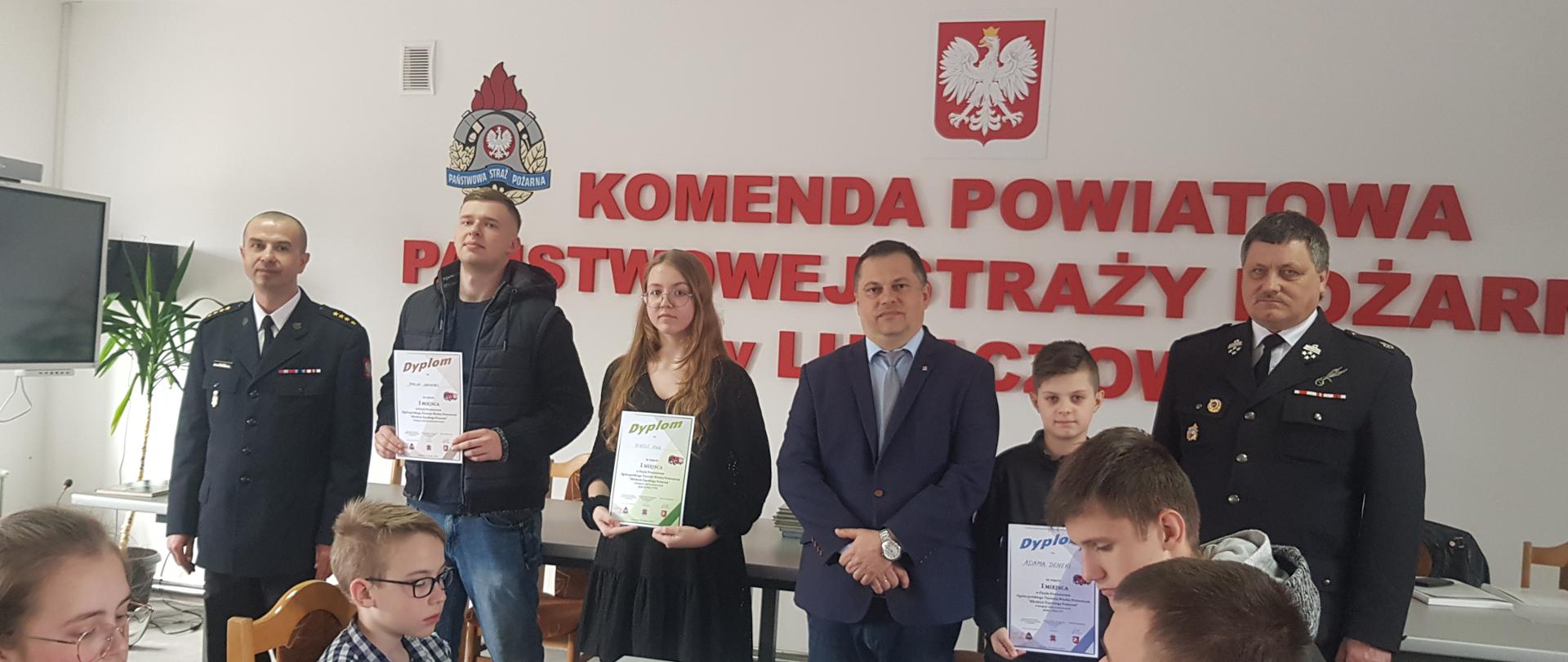 Zdjęcie przedstawia laureatów trzech pierwszych miejsc w każdej grupie wiekowej OTWP w 2022, wraz z przybyłymi gośćmi. Na ścianie napis Komenda Powiatowa Państwowej Straży Pożarnej w Lubaczowie