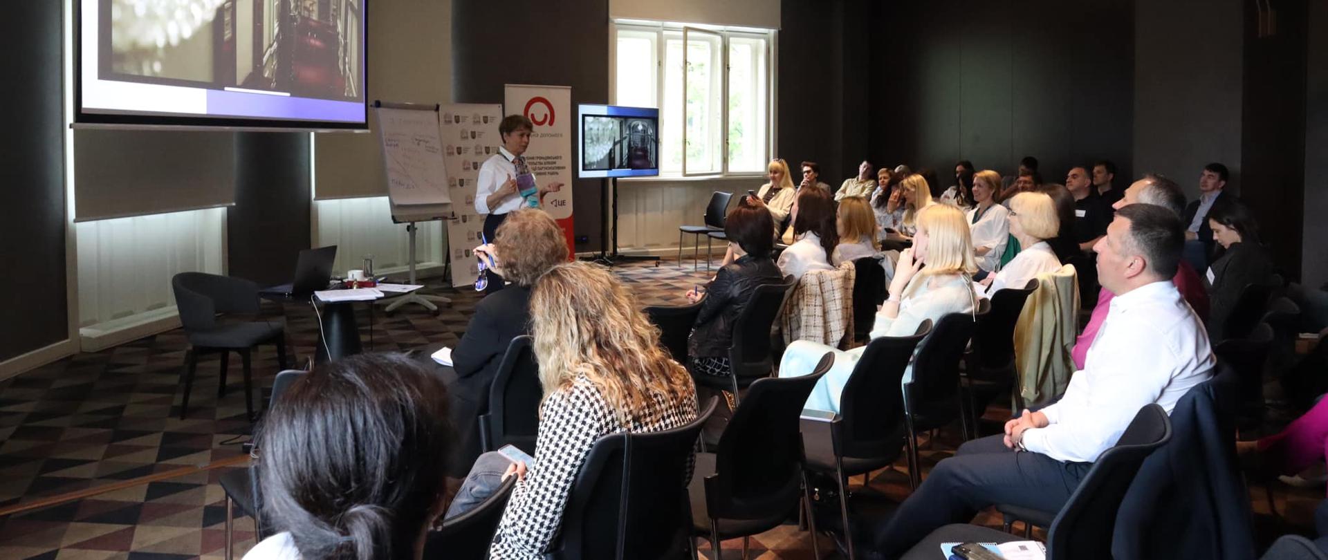 Sala konferencyjna, na krzesłach siedzą uczestnicy spotkania, przed nimi stoi kobieta prowadząca szkolenie, a za nią baner z logo Polska pomoc