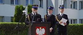 trzech strażaków ubranych w mundury galowe stoi przed amboną, jeden trzyma w dłoniach dokumenty, drugi przygląda się pierwszemu, który przemawia do gości, w tle budynek komendy straży pożarnej, żywopłot oraz dwa rosnące drzewa, pogoda słoneczna