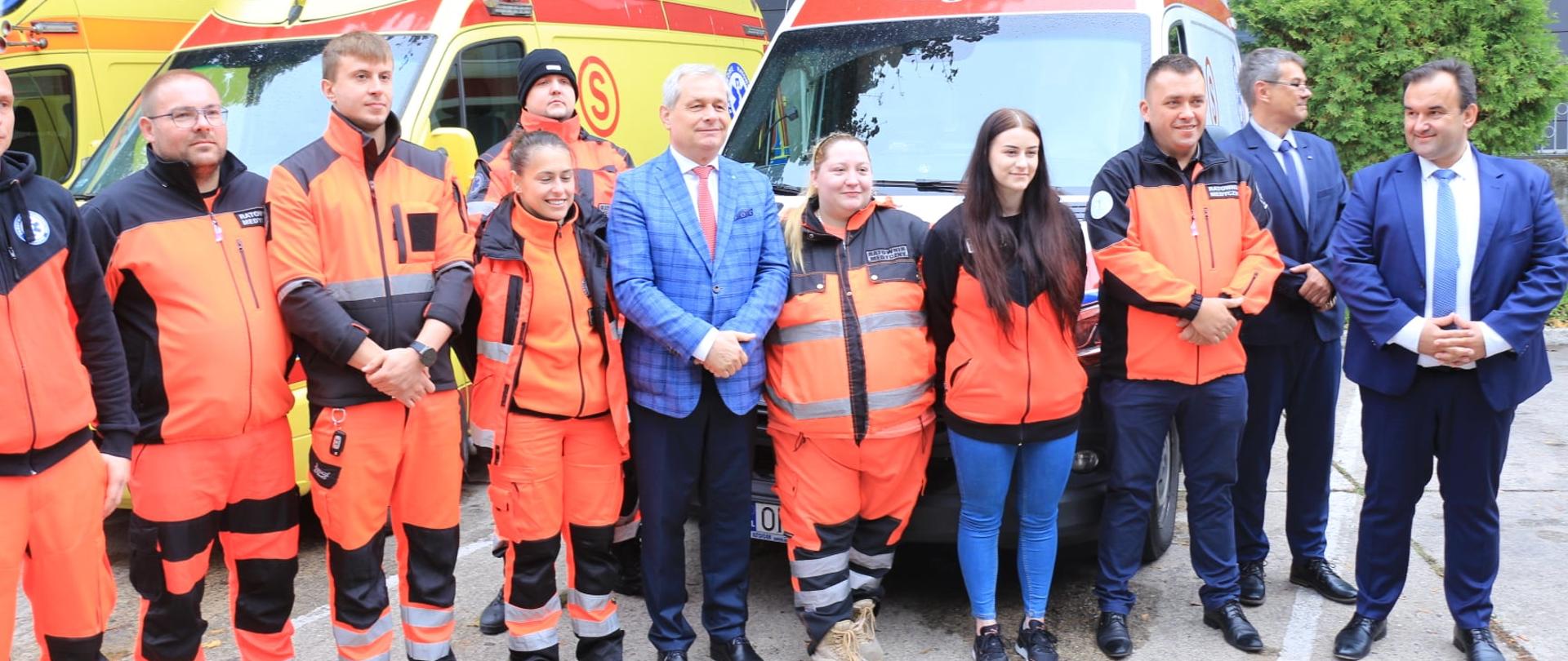 Na zdjęciu zespół ratowników medycznych, ubranych w pomarańczowe stroje. Grupa ratowników pozujących do zdjęcia wspólnie z wojewodą opolskim i dyrekcją OCRM w Opolu. Na zdjęciu łącznie 10 osób, stojących obok siebie. W grupie tej wyróżnia się wojewoda opolski, który ubrany jest w niebieską marynarkę w kratkę. W tle karetki. 