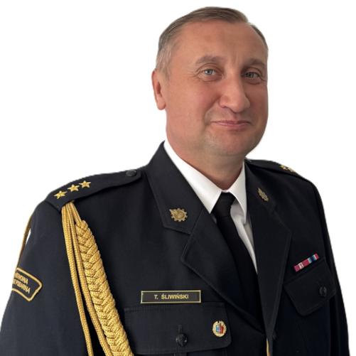 Zdjęcie przedstawia mężczyznę w formalnym mundurze służbowym. Ma krótkie, jasne włosy i poważny wyraz twarzy. Mundur jest granatowy, z krawatem, ozdobnymi złotymi sznurami na ramieniu oraz odznaczeniami i naszywkami na piersi. Tło jest jasne i jednolite.