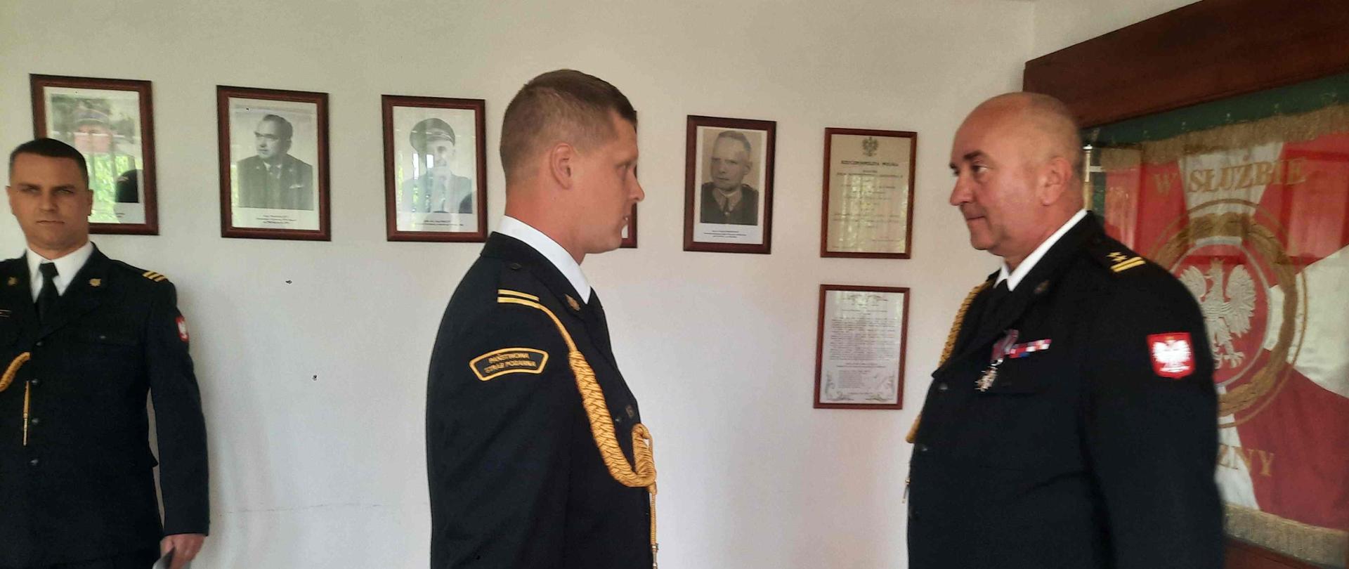 Na zdjęciu dwóch strażaków w umundurowaniu galowym stoi naprzeciw siebie. W tle na ścianach sztandar Komendy zdjęcia i dokumenty w oprawach.