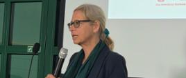 Na zdjęciu widoczna jest kobieta przemawiająca przy mównicy podczas konferencji. W dłoni trzyma mikrofon, a za nią znajduje się ekran z wyświetloną prezentacją zawierającą logotypy i informacje o partnerach wydarzenia. Fotografia została wykonana we wnętrzu sali konferencyjnej.