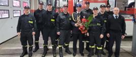 Strażacy stoją na tle drabiny pożarniczej z kończącym służbę Tadeuszem Wieczorkiem. Tadeusz trzyma w rękach statuetkę pamiątkową, gaśnicę okolicznościową i kwiaty.