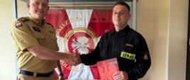 Na pierwszym planie Komendant Powiatowy Państwowej Straży Pożarnej w Rypinie bryg. Piotr Wypych oraz sekc. Dawid Dąbrowski podczas wręczania aktu mianowania na stanowisko ratownika. Strażacy podają sobie dłonie. W tle widoczny jest sztandar Komendy Powiatowej Państwowej Straży Pożarnej w Rypinie.a