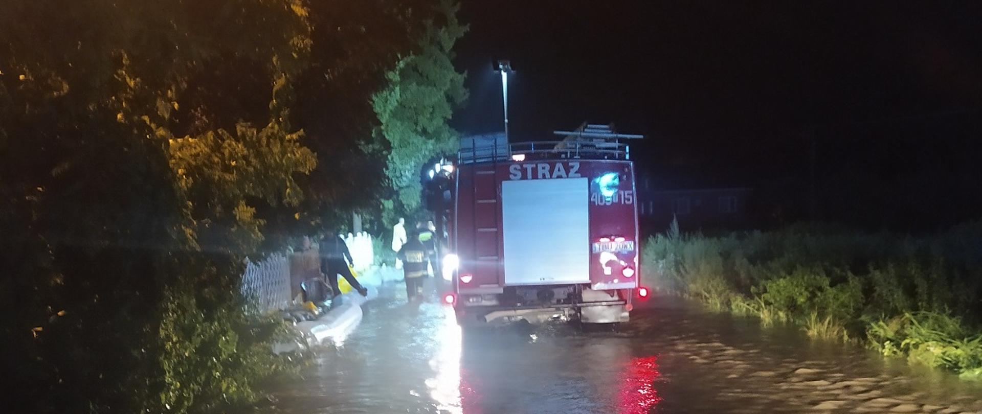 Zdjęcie przedstawia samochód pożarniczy stojący na częściowo zalanej drodze, który oświetla teren działania