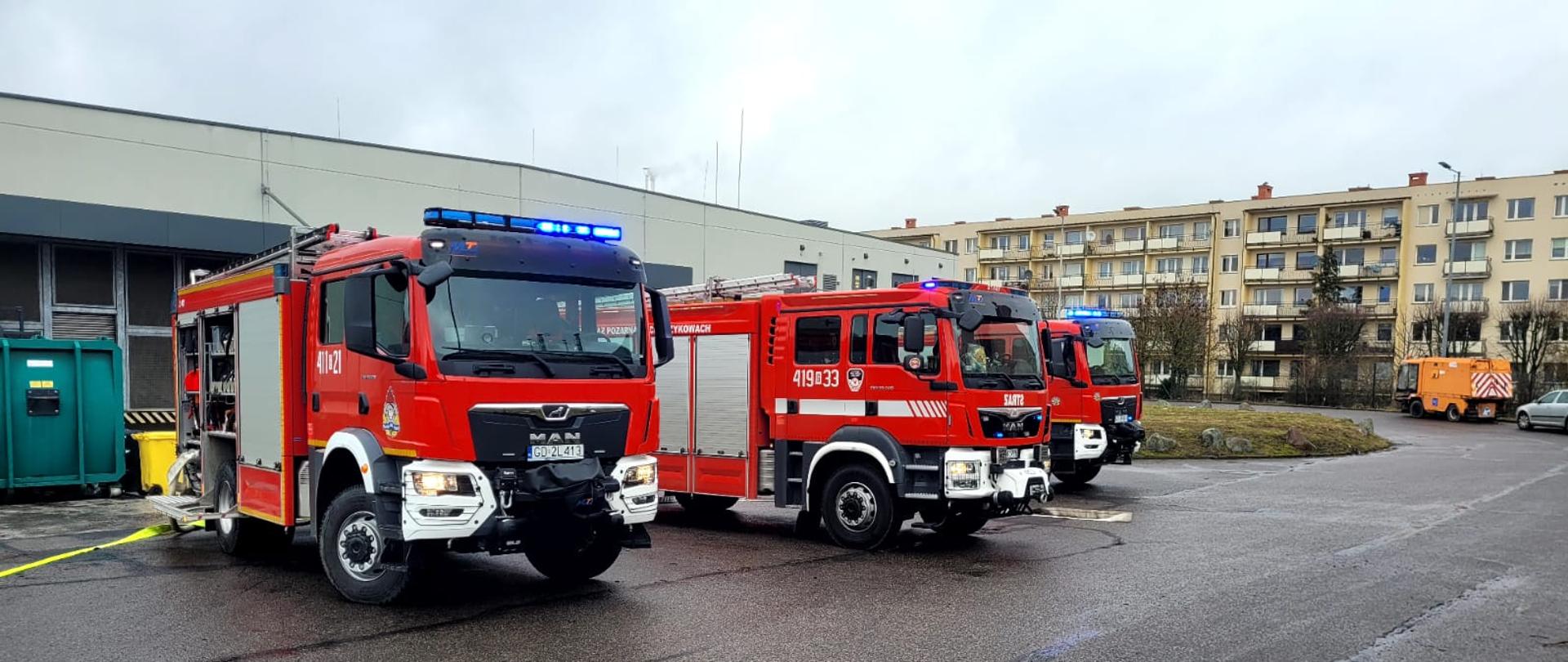 Pochmurny dzień na mokrym czarnym asfalcie ustawione w rzędzie trzy czerwone samochody strażackie na tle magazynu obiektu usługowo handlowego i architektury miejskiej. Wszystkie wozy bojowe mają włączone światła mijania oraz niebieskie oświetlenie ostrzegawcze. 
