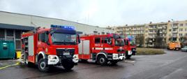 Pochmurny dzień na mokrym czarnym asfalcie ustawione w rzędzie trzy czerwone samochody strażackie na tle magazynu obiektu usługowo handlowego i architektury miejskiej. Wszystkie wozy bojowe mają włączone światła mijania oraz niebieskie oświetlenie ostrzegawcze. 