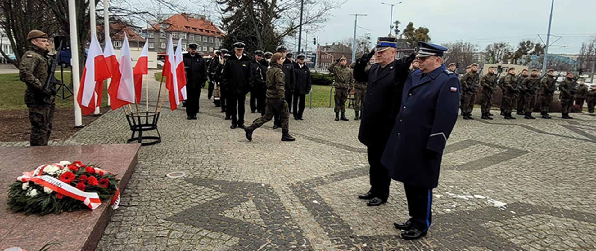 Funkcjonariusze Państwowej Straży Pożarnej oraz Policji stojąc obok siebie oddają honor obok stoją przedstawiciele Sił Zbrojnych Polski. 