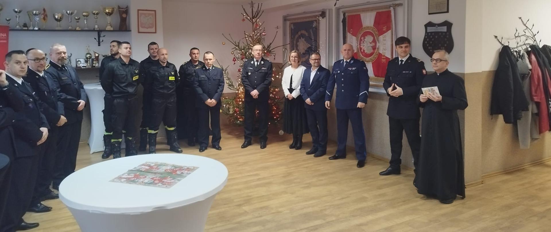 Zdjęcie przedstawia księdza, funkcjonariuszy straży pożarnej oraz zaproszonych gości na spotkaniu wigilijnym.