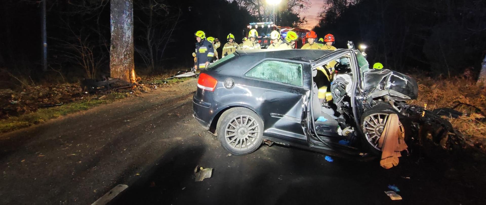 Na pierwszym planie mocno rozbity samochód osobowy, dalej strażacy oraz wóz strażacki oświetlający miejsce zdarzenia.
