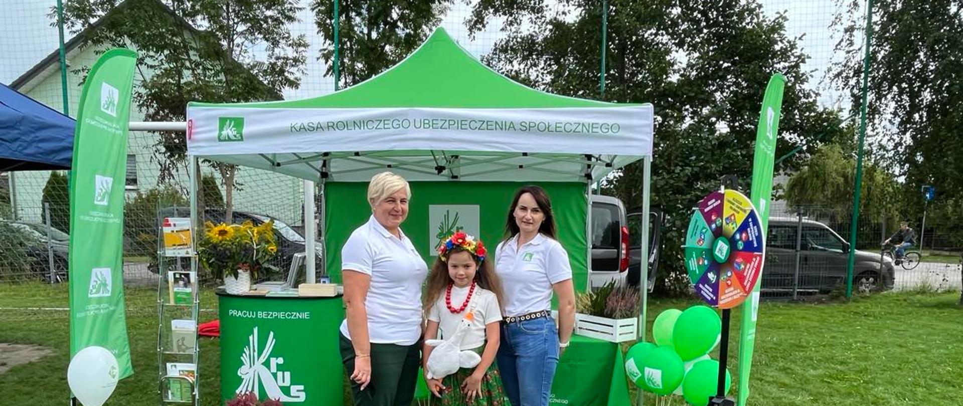 Na tle zielonego namiotu ze stoiskiem informacyjnym KRUS stoją dwie kobiety pracownice KRUS z dziewczynką w stroju ludowym. Obok zielone balony i gra prewencyjna Koło Fortuny