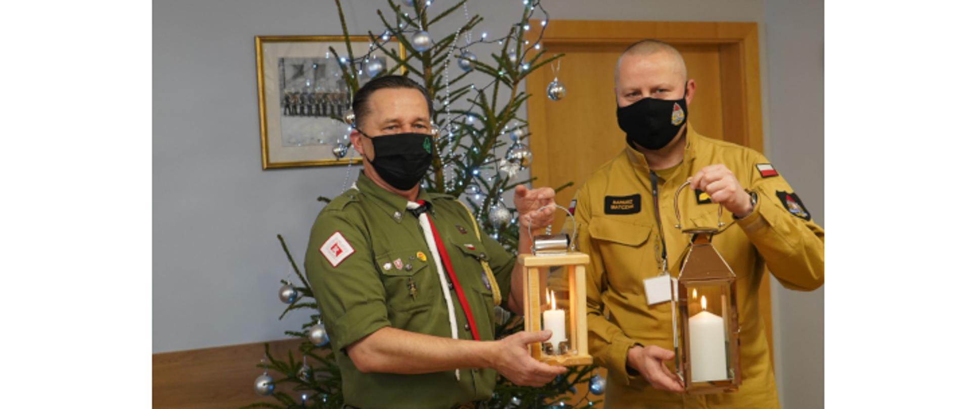 mężczyzna harcerz tzrymający w reku drewniany lampion z paląca się świeczką, obok stoi strażak z metalowym lampionem w środku z palacą się świeczką, w tle choinka 