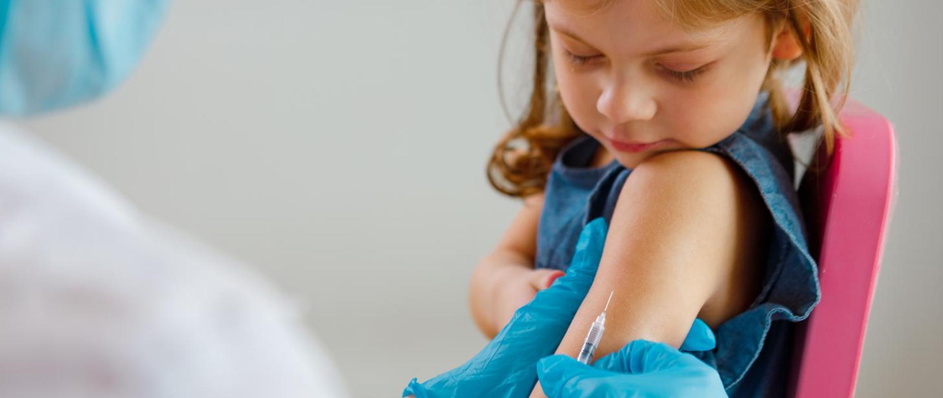 Zdjęcia przedstawia dziewczynkę której podawana jest szczepionka w lewę ramie. 