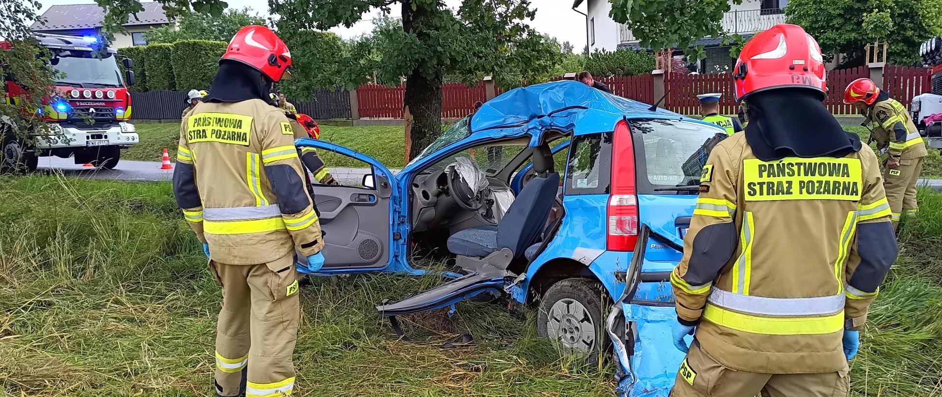 Na pierwszym planie dwóch strażaków w ubraniach specjalnych i hełmach. Na środku niebieski samochód osobowy po wypadku. Ma silnie odkształconą lewą stronę. Widoczne ślady usuwania drzwi i słupka. W tle uderzone drzewo, droga i samochody strażackie po bokach.