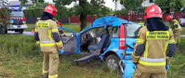Na pierwszym planie dwóch strażaków w ubraniach specjalnych i hełmach. Na środku niebieski samochód osobowy po wypadku. Ma silnie odkształconą lewą stronę. Widoczne ślady usuwania drzwi i słupka. W tle uderzone drzewo, droga i samochody strażackie po bokach.