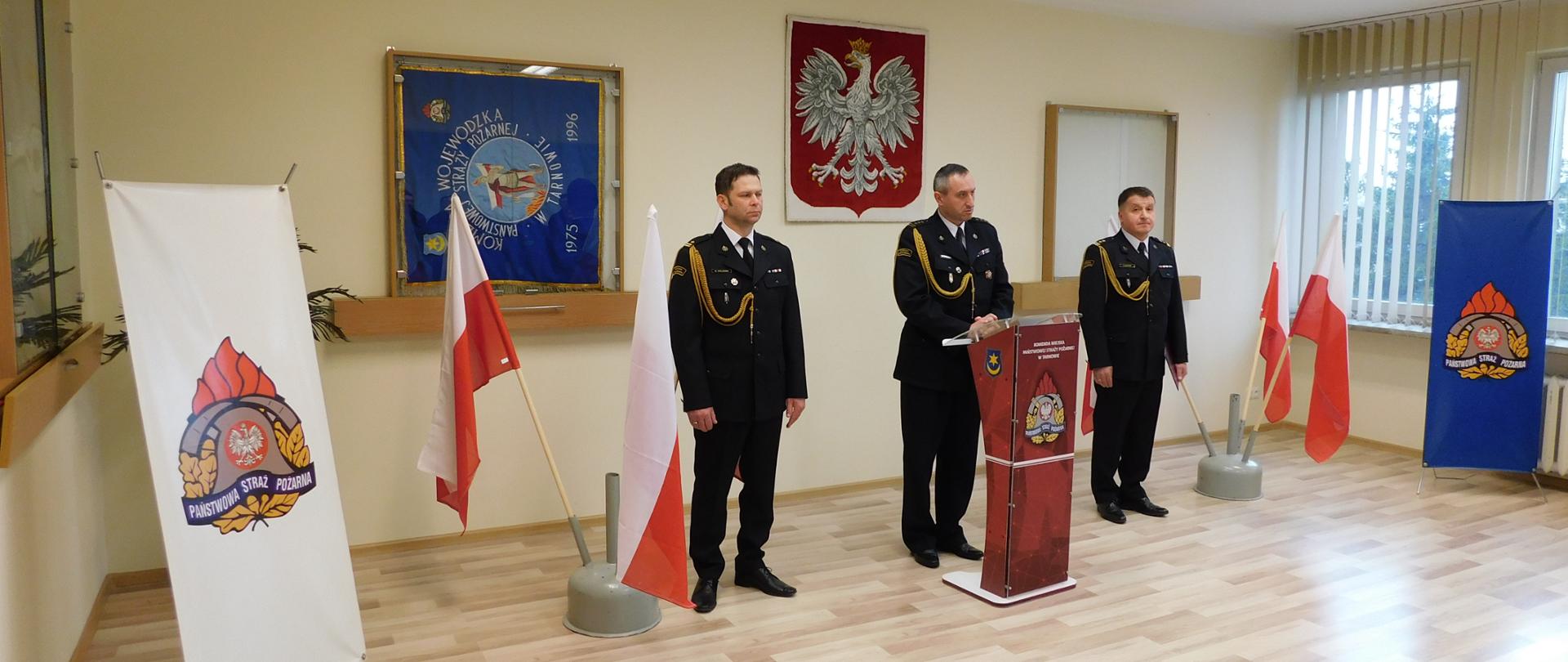 Na zdjęciu widoczni są 3 funkcjonariusze podczas przemówienia z prawej i lewej strony widoczne są flagi państwowe oraz loga PSP. Przemówienie ma miejsce na Sali narad KM PSP w Tarnowie.