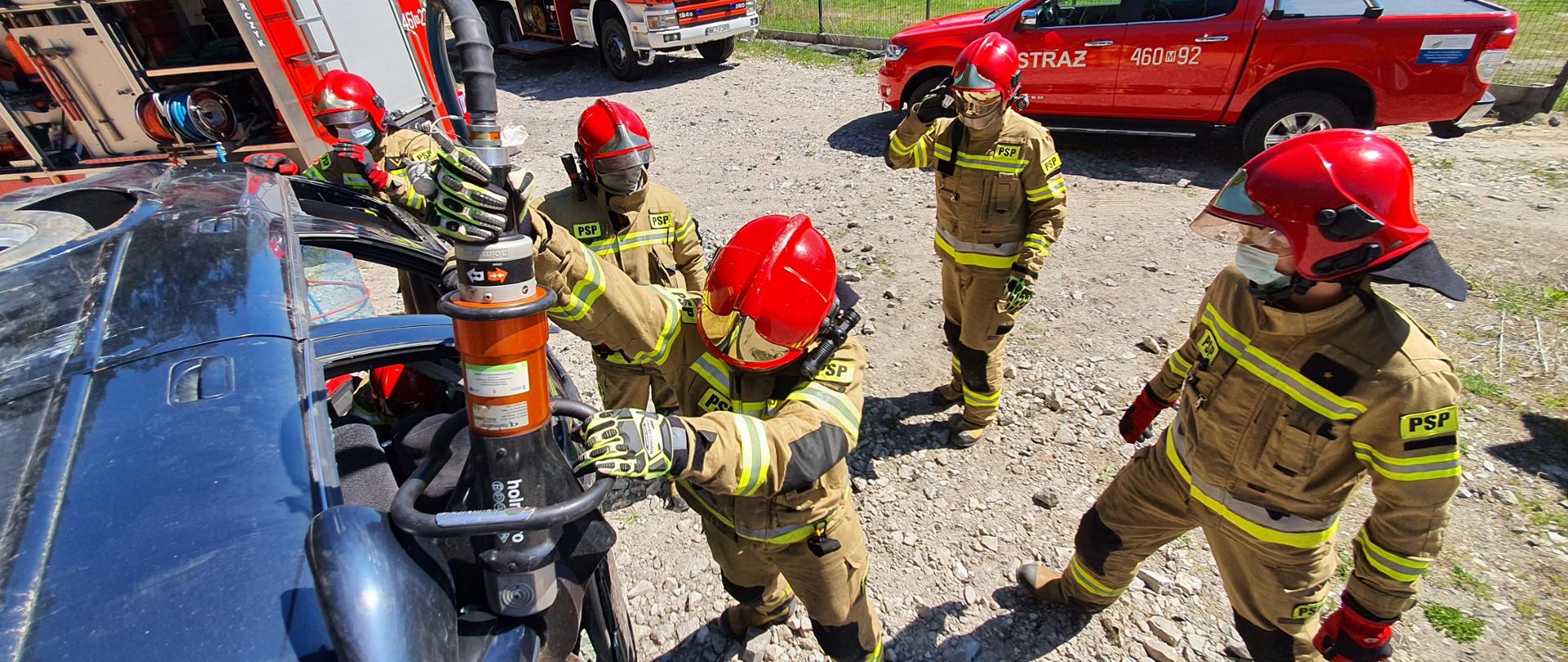 Strażacy w umundurowaniu specjalnym podczas ćwiczeń używają nożyc hydraulicznych do cięcia karoserii samochodu