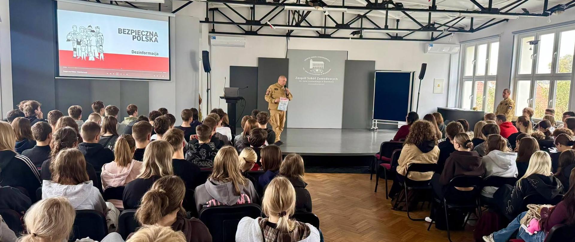Na fotografii widoczne jest spotkanie edukacyjne z młodzieżą poświęcone tematyce bezpieczeństwa i przeciwdziałania dezinformacji. Strażak prowadzi prelekcję dla licznie zgromadzonych uczniów, wykorzystując materiały programu „Bezpieczna Polska”. Spotkanie miało na celu zwiększenie świadomości młodzieży w zakresie rozpoznawania zagrożeń informacyjnych oraz właściwego reagowania na fałszywe lub niezweryfikowane informacje.