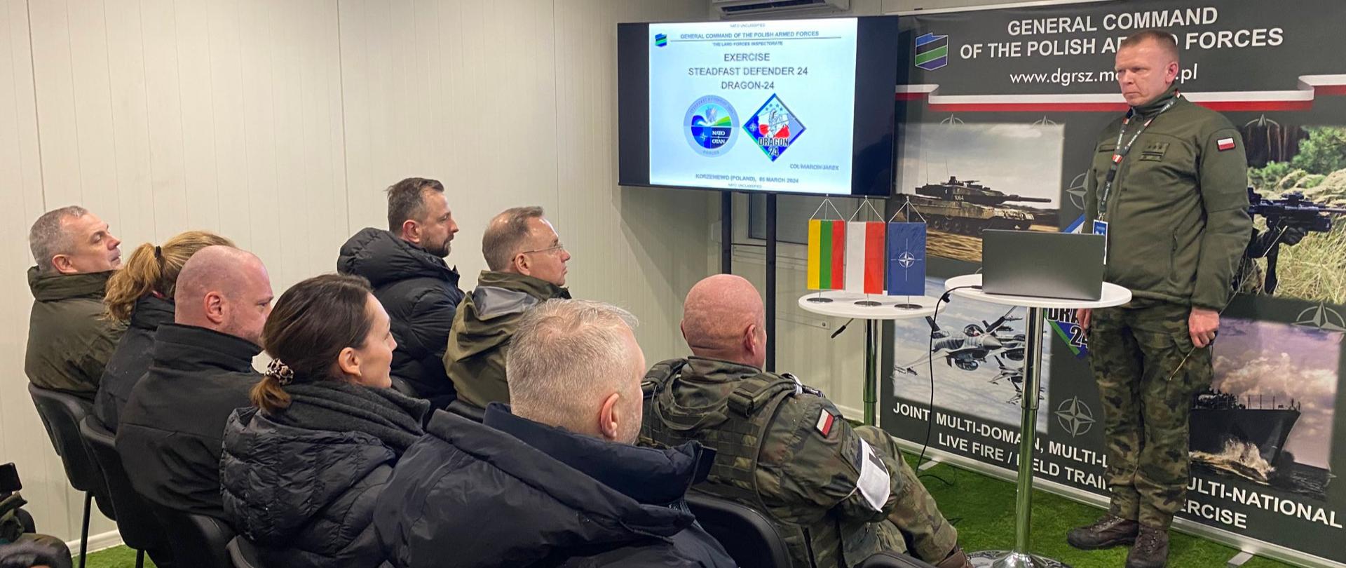kilka osób siedzi w sali, przed nimi stoi żołnierz, za nim tablica ze zdjęciami z manewrów 