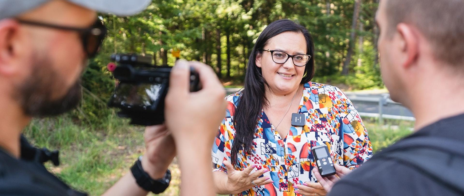Wiceminister Małgorzata Jarosińska-Jedynak stoi przy drodze w lesie i rozmawia z dziennikarzami