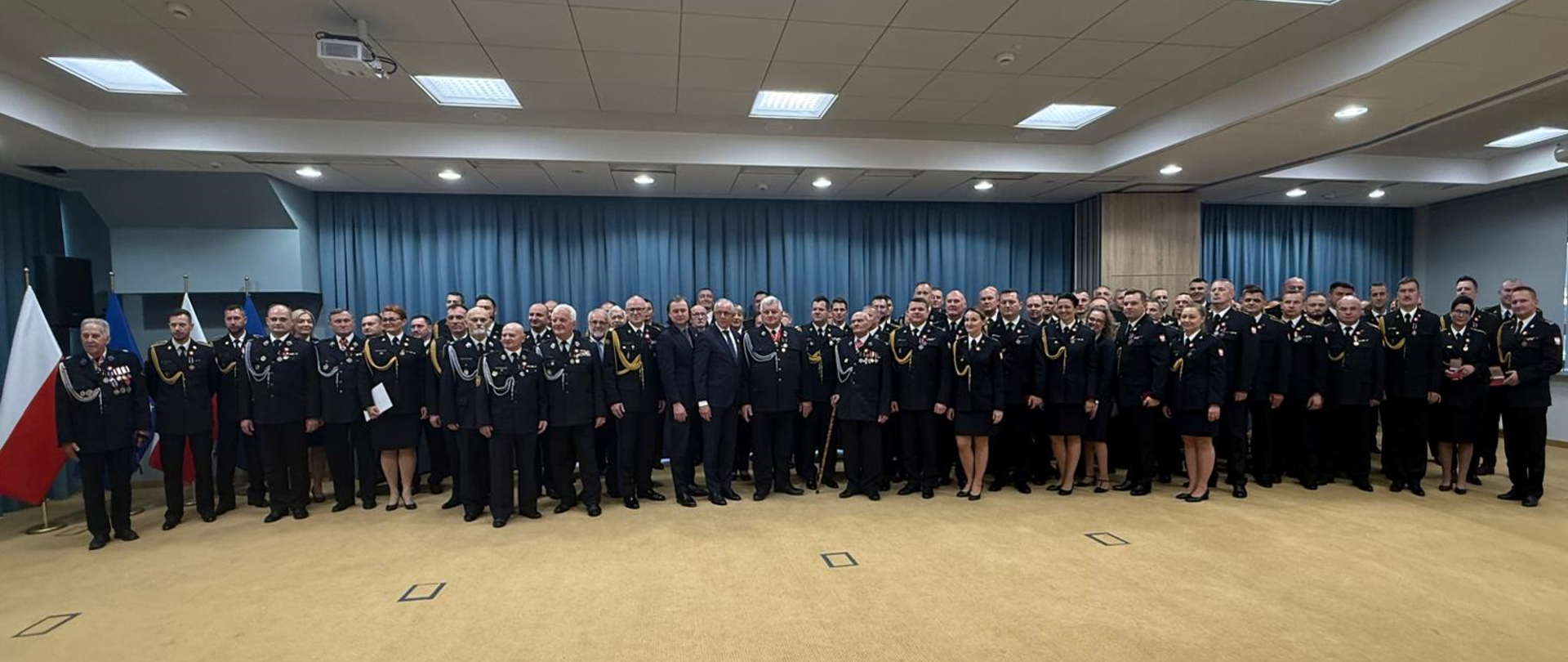 Zdjęcie panoramiczne przedstawiające dużą grupę funkcjonariuszy PSP w galowych mundurach ustawionych w kilku rzędach podczas uroczystości. W tle znajdują się zasłony oraz flagi Polski i Unii Europejskiej. Uczestnicy stoją na jasnej wykładzinie w sali konferencyjnej.