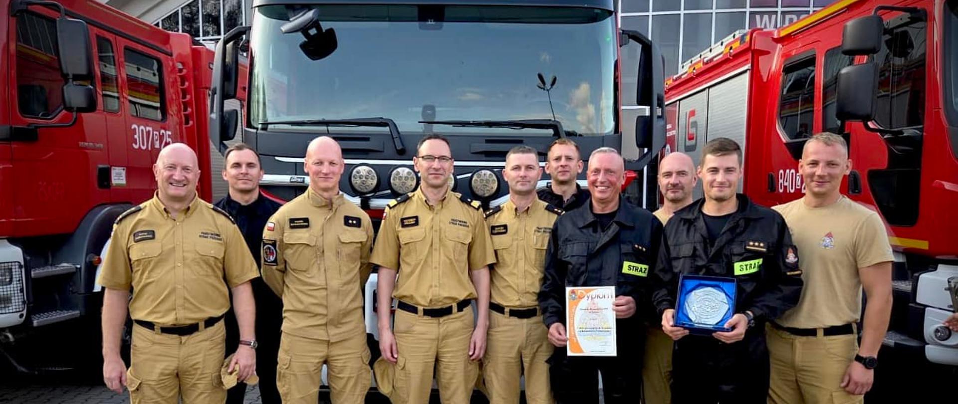 Grupa dziesięciu strażaków w mundurach służbowych, stoją przed ekspozycją aut pożarniczych. Dwóch z nich trzyma trofeum zawodów - jeden dyplom, drugi okolicznościową statuetkę. 
