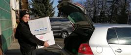 Widok z boku. Członek OSP stoi za samochodem osobowym i trzyma w rękach paczki z maseczkami ochronnymi. 