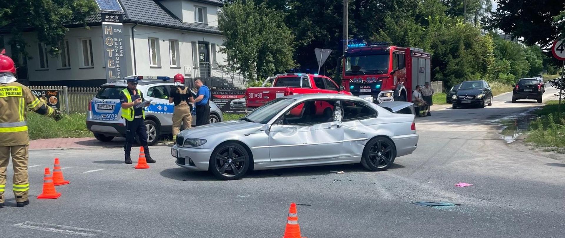 Zdjęcie przedstawia drogę wojewódzką: na pierwszym planie stoi zniszczony samochód osobowy, a za nim w tle pojazdy służb ratowniczych: radiowóz i dwa samochody pożarnicze. 